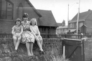 Janet Brown and her cousins sitting on the wall around Manor Farm, with the Village Hall behind, mid-1960s.