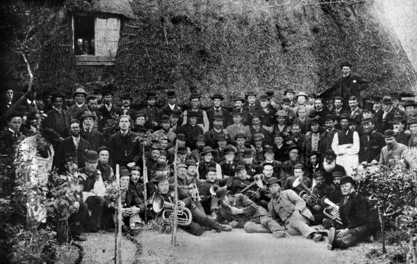 A group of nearly 100 men gathered in front of a thatched cottage, with the Trumpington Brass Band in foreground. The photograph has been dated between the 1860s and the 1880s (probably early 1880s). Percy Robinson collection.
