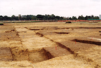 One of three Neolithic burial monuments being excavated on the Trumpington Meadows site, with burials marked by white posts, burials in rectangular area, blue pegs marking earlier inner ring, red pegs outer ring with pottery dating from c. 3500BC, with Trumpington Church and Anstey hall in the distance. Photo: Andrew Roberts, 3 October 2010