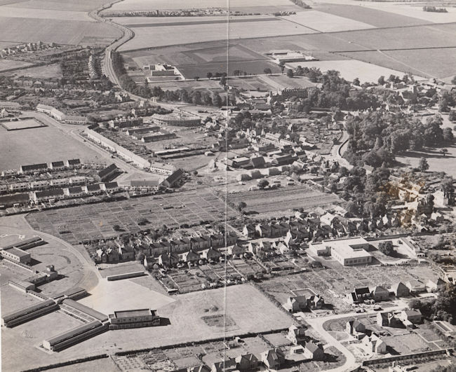 Aerial view of Trumpington from the north east, 1955. Includes the village core, High Street, Church Lane, Shelford Road, Bishop's Road, Hauxton Road, Anstey Hall, Church, PBI headquarters building and grounds, Cambridge-Bedford railway line, Anstey Hall Farm shepherd's cottage and hay stacks, former PoW Camp, the Estate and King George V recreation ground (with bowling green), Anstey Way with shops under construction, Alpha Terrace, Fawcett School, allotments, Wingate Way under construction, The Red Lion, Manor Farm with hay ricks, Village Hall, blacksmiths. Cambridgeshire Collection.